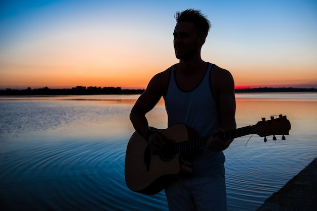 Siluetta di giovane uomo bello che gioca chitarra in riva al mare durante l'alba