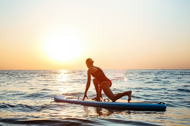 Siluetta di yoga di pratica della bella donna sulla tavola da surf ad alba.