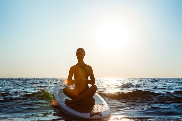 Siluetta di yoga di pratica della bella donna sulla tavola da surf ad alba.