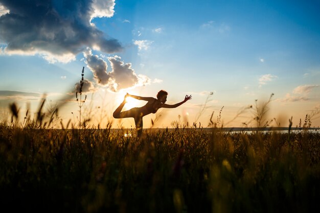 Siluetta di yoga di pratica della ragazza allegra nel campo ad alba.