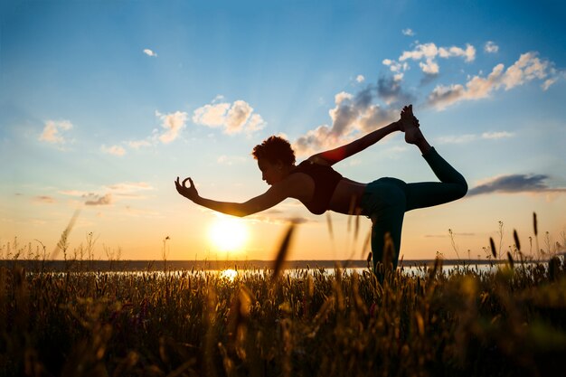 Siluetta di yoga di pratica della ragazza allegra nel campo ad alba.