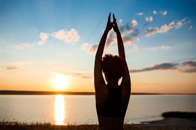 Siluetta di yoga di pratica della ragazza allegra nel campo ad alba.