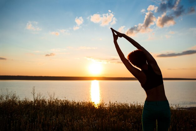 Siluetta di yoga di pratica della ragazza allegra nel campo ad alba.