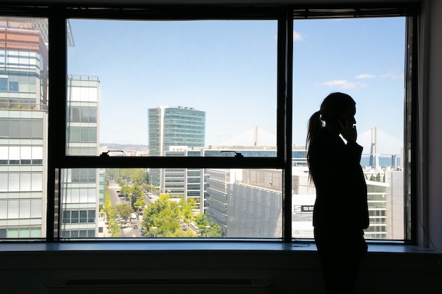 Siluetta irriconoscibile della donna di affari che sta vicino alla finestra e che parla sul telefono cellulare. Manager professionista in ombra e paesaggio urbano. Business, comunicazione e concetto di azienda