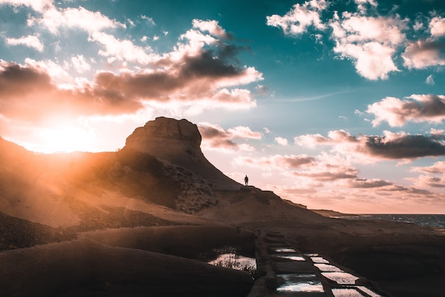 Siluetta umana che sta su una montagna rocciosa durante il tramonto sotto un cielo blu nuvoloso