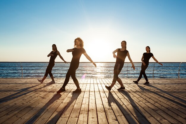 Siluette delle donne allegre che ballano zumba vicino al mare all'alba