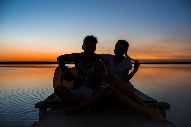 Siluette di giovani belle coppie che riposano rallegrandosi all'alba vicino al lago