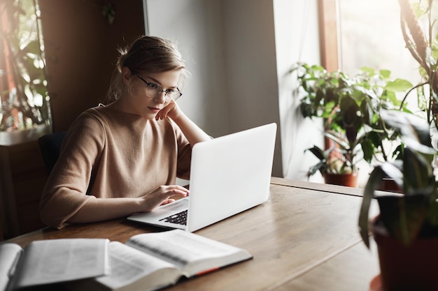 Simpatica collega europea con gli occhiali seduta vicino alla finestra nel caffè che si appoggia la testa a portata di mano e legge articoli in rete tramite laptop alla ricerca di informazioni per lavoro prendendo appunti sul taccuino