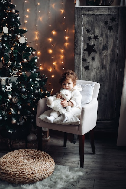 Simpatico bambino riccio con un orso di peluche seduto in poltrona vicino a un albero di Natale splendidamente decorato