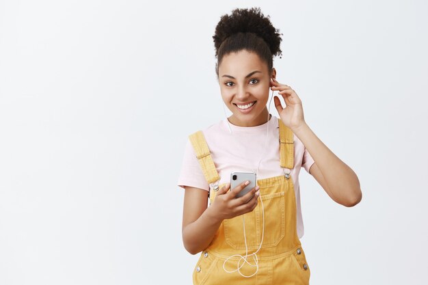 Sintonizzati su grandi vibrazioni. bella donna afroamericana felice in tuta gialla, indossare gli auricolari, tenere in mano lo smartphone, scegliere una canzone per uscire e camminare per le strade della città