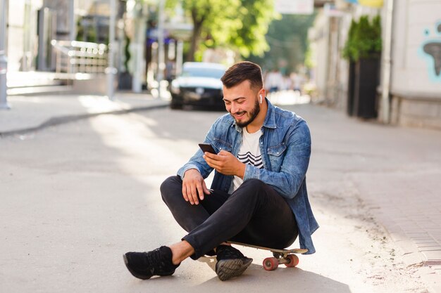 Skateboarder maschio che si siede sul pattino facendo uso del telefono cellulare