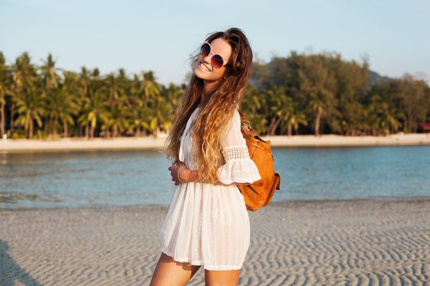 Slim bella donna in abito bianco sulla spiaggia tropicale sul tramonto che tiene lo zaino in pelle.