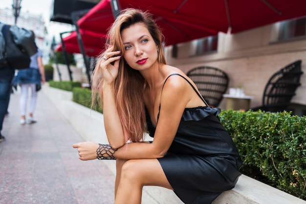 Slim donna abbronzata in elegante abito nero e tacchi con i capelli biondi luminosi in posa nella vecchia città europea vicino al ristorante di lusso.