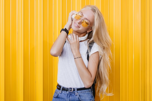 Slim donna sognante con lunghi capelli lucidi godendo buona giornata. Ritratto di bella ragazza abbronzata in maglietta bianca in posa su sfondo giallo.