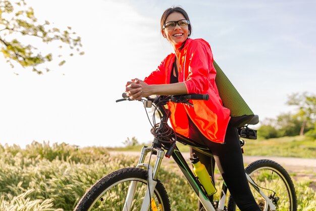 Slim fit bella donna che fa sport mattina nel parco in sella alla bicicletta con materassino yoga in abito fitness colorato, esplorando la natura, sorridendo felice stile di vita sano