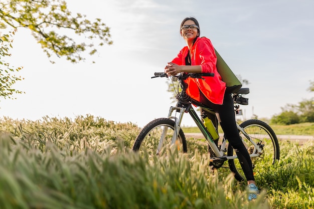 Slim fit bella donna che fa sport mattina nel parco in sella alla bicicletta con materassino yoga in abito fitness colorato, esplorando la natura, sorridendo felice stile di vita sano