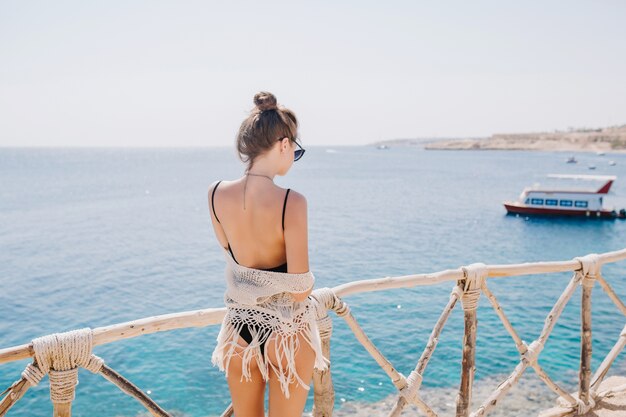 Slim ragazza graziosa con acconciatura alla moda che gode della vista sul mare e guardando il lancio. Ritratto dal retro della giovane donna incredibile in costume da bagno nero in piedi sull'oceano