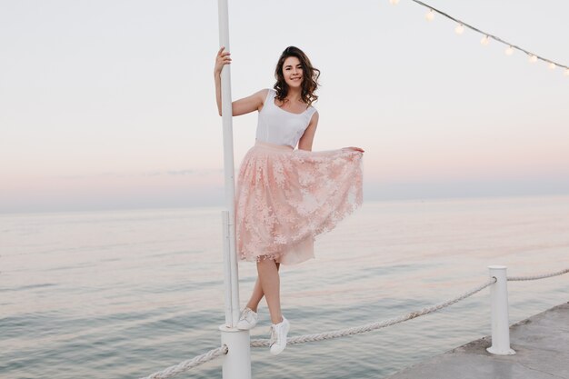 Slim ragazza in gonna rosa e scarpe bianche in posa con le gambe incrociate e un sorriso ispiratore sul mare