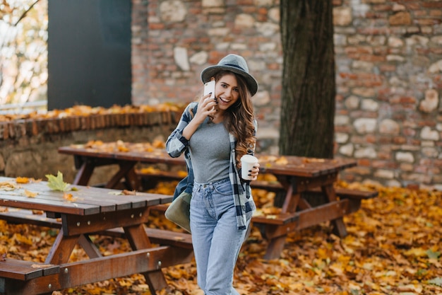 Slim ragazza in pantaloni denim blu parlando al telefono nel parco con alberi dietro