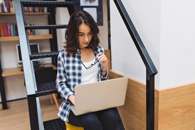 Smart carino giovane donna seduta sulle scale in libreria, lavorando con il computer portatile. Studio, studente intelligente, navigazione in internet, vita universitaria, libero professionista, start-up, reportage.