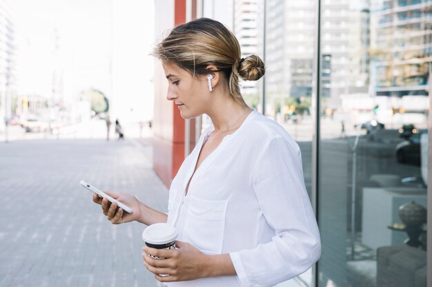 Smart Phone biondo della tenuta della giovane donna e tazza di caffè asportabile in mani