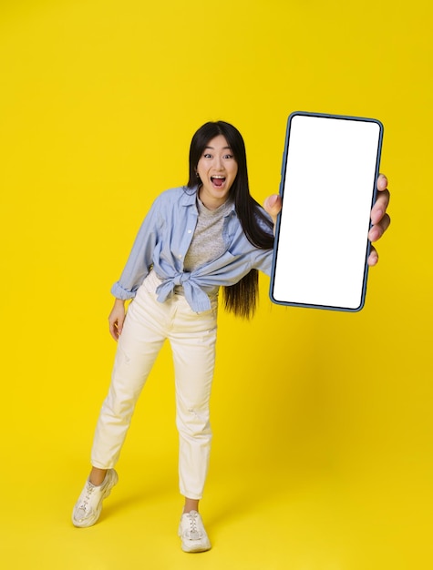 Smartphone felice della tenuta della giovane ragazza asiatica che mostra la pubblicità dell'app mobile dello schermo verde e il sorriso eccitato sulla macchina fotografica isolata su fondo giallo Grande offerta Posizionamento del prodotto