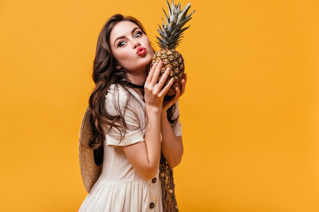 Snella ragazza dagli occhi verdi in abito bianco soffia un bacio e tiene l'ananas.