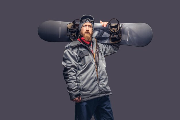 Snowboarder rosso brutale con una barba piena in un cappello invernale e occhiali protettivi vestito con un cappotto da snowboard in posa con uno snowboard in uno studio, guardando una telecamera. Isolato su uno sfondo grigio
