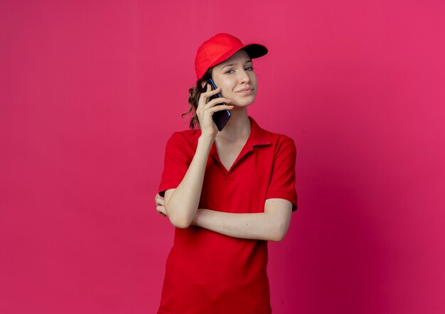 Soddisfatta giovane bella ragazza delle consegne in uniforme rossa e cappuccio in piedi con la postura chiusa parlando al telefono guardando la telecamera isolata su sfondo cremisi con spazio di copia