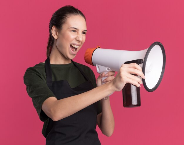 Soddisfatta giovane ragazza bruna barbiere in uniforme tiene altoparlante e flacone spray guardando a lato sul rosa