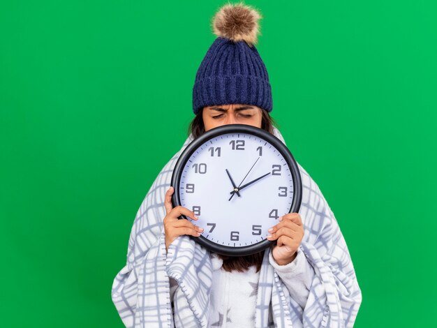 Soddisfatta giovane ragazza malata con gli occhi chiusi che indossa cappello invernale con sciarpa viso coperto con orologio da parete isolato su sfondo verde