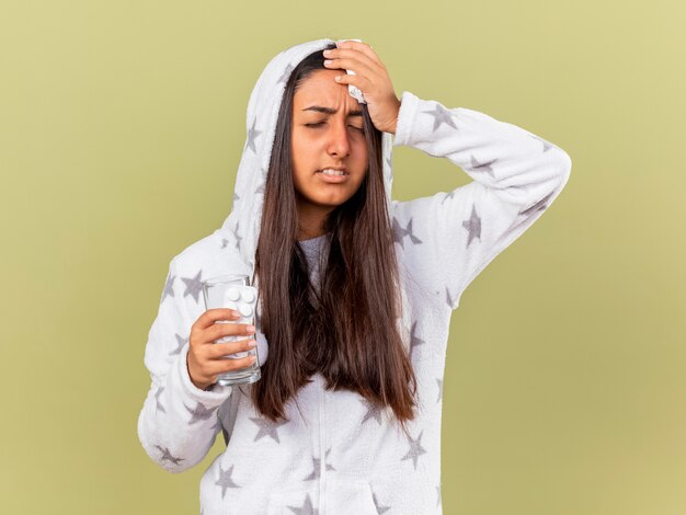 Soddisfatta giovane ragazza malata con gli occhi chiusi che mette sul cofano tenendo il bicchiere di acqua con le pillole e mettendo la mano sulla fronte isolata su sfondo verde oliva