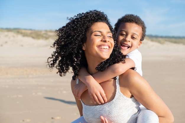 Soddisfatti madre e figlio che trascorrono del tempo sulla spiaggia. Famiglia afroamericana che cammina, ride, gioca, cavalca. Tempo libero, tempo in famiglia, concetto di genitorialità