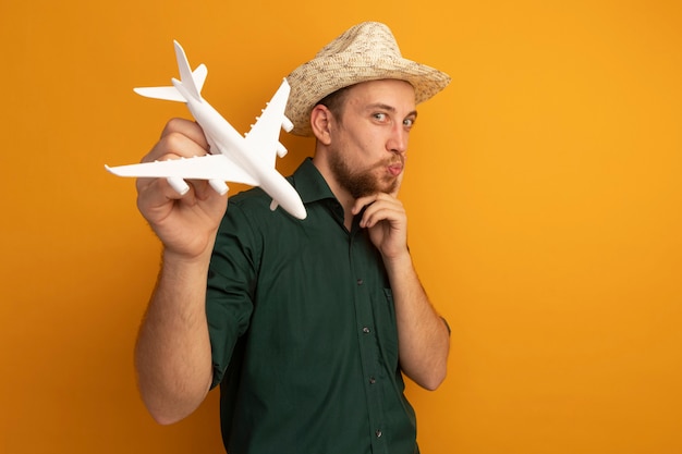 Soddisfatto bell'uomo biondo con cappello da spiaggia mette il dito sul viso e tiene l'aereo modello sull'arancia