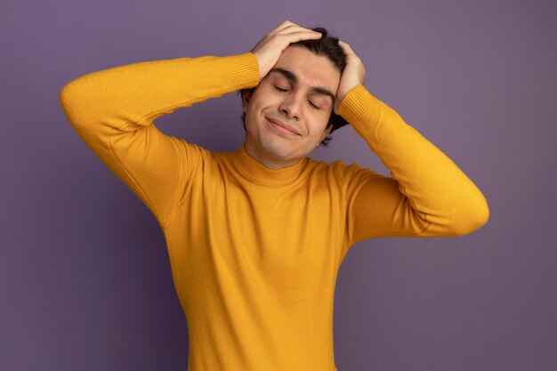 Soddisfatto con gli occhi chiusi giovane bel ragazzo che indossa un maglione dolcevita giallo che mette le mani sulla testa isolata sul muro viola