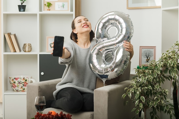 Soddisfatto degli occhi chiusi bella donna nel giorno delle donne felici che tiene il palloncino numero otto con il telefono seduto sulla poltrona nel soggiorno