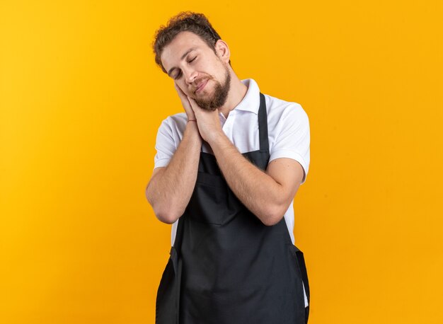 Soddisfatto degli occhi chiusi, giovane barbiere maschio che indossa l'uniforme che mostra il gesto del sonno isolato sul muro giallo