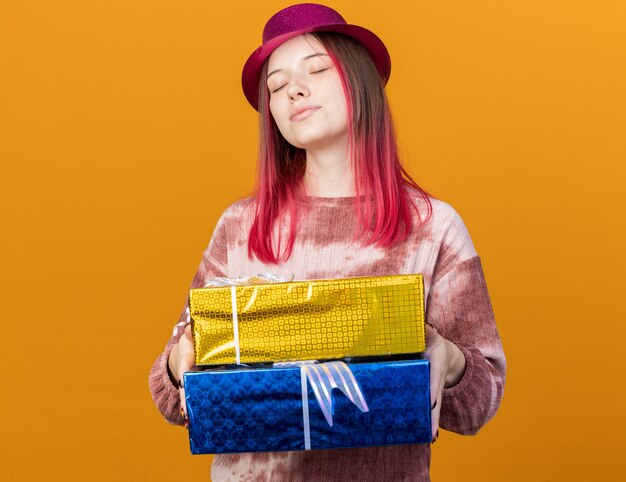 Soddisfatto degli occhi chiusi, giovane e bella ragazza che indossa un cappello da festa che tiene in mano scatole regalo