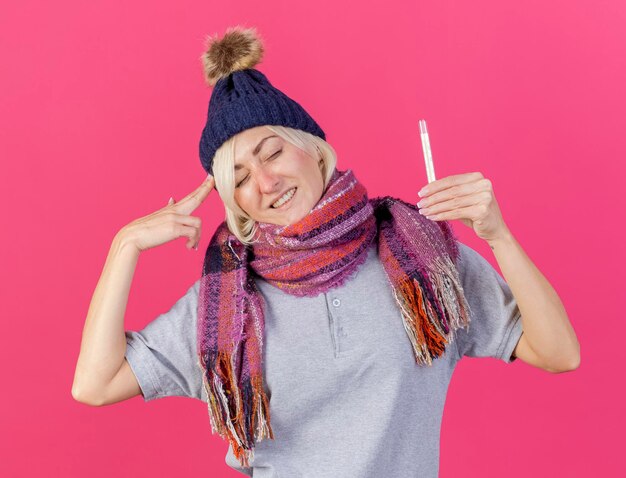Soddisfatto giovane bionda malata donna slava che indossa il cappello invernale e gesti sciarpa segno della mano della pistola