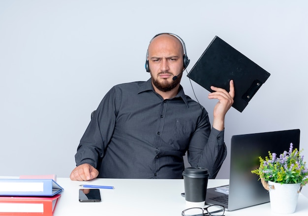 Soddisfatto giovane calvo call center uomo che indossa auricolare seduto alla scrivania con strumenti di lavoro che tengono appunti isolati su sfondo bianco