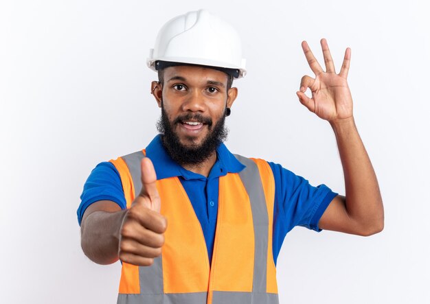 Soddisfatto giovane costruttore afro-americano uomo in uniforme con casco di sicurezza sfogliando e gesticolando segno ok isolato su sfondo bianco con spazio di copia