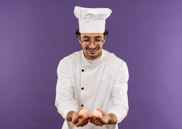 Soddisfatto giovane cuoco maschio che indossa l'uniforme dello chef e bicchieri tenendo e guardando le uova sulla porpora