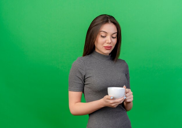 Soddisfatto giovane donna graziosa che tiene e guardando la tazza isolato su sfondo verde con spazio di copia
