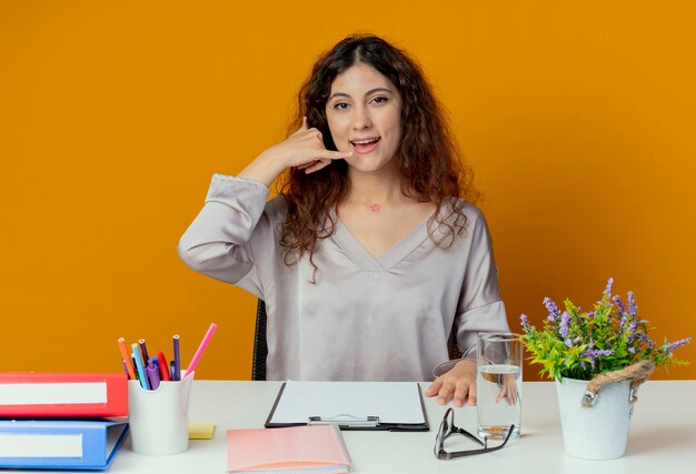 Soddisfatto giovane impiegato abbastanza femminile che si siede allo scrittorio con gli strumenti dell'ufficio che mostrano il gesto di telefonata isolato sull'arancio