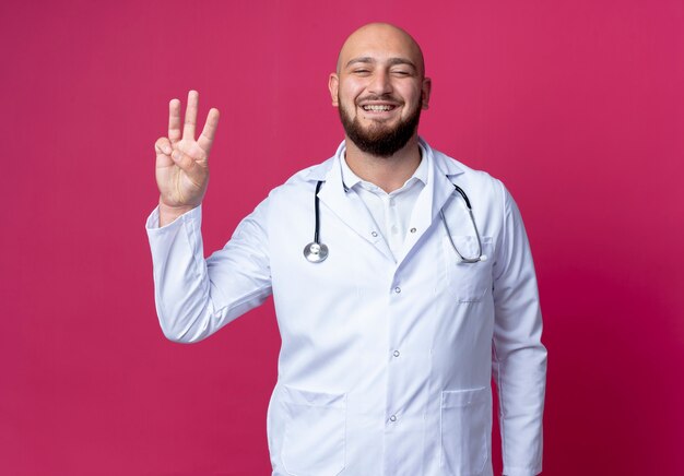 Soddisfatto giovane medico maschio calvo che indossa veste medica e stetoscopio che mostra tre isolati sulla parete rosa