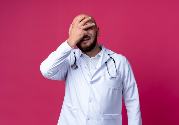 Soddisfatto giovane medico maschio che indossa abito medico e stetoscopio ha coperto il viso