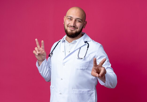 Soddisfatto giovane medico maschio che indossa veste medica e stetoscopio che mostra gesti okey isolati sul rosa