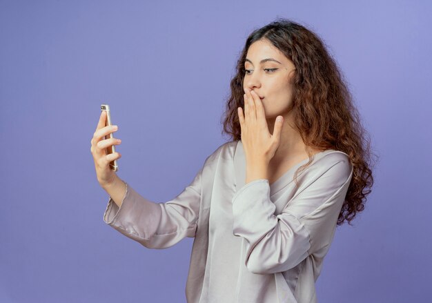 Soddisfatto giovane ragazza graziosa che tiene e che guarda il telefono e la bocca coperta con la mano isolata sulla parete blu