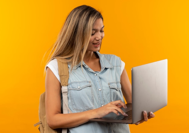 Soddisfatto giovane ragazza graziosa dell'allievo che indossa la borsa posteriore utilizzando il computer portatile isolato sulla parete arancione