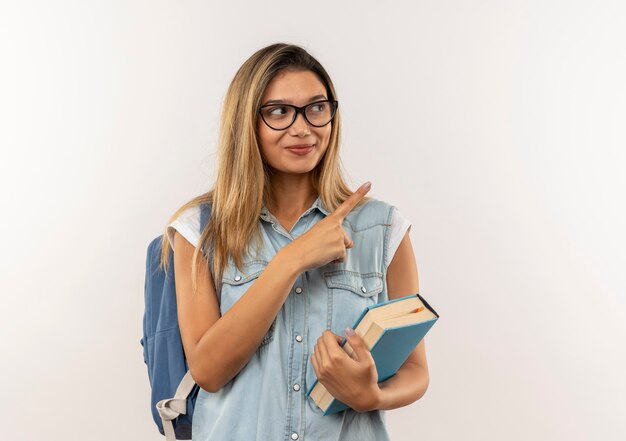 Soddisfatto giovane ragazza graziosa dell'allievo con gli occhiali e la borsa posteriore che tiene il libro che indica e che esamina il lato isolato sul muro bianco